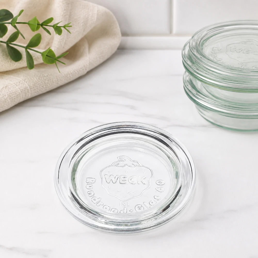 Coperchio del vaso WECK RR60 Coperchio in vetro WECK RR60 vista impilata di diversi coperchi sovrapposti su marmo con telo da cucina e ramo verde