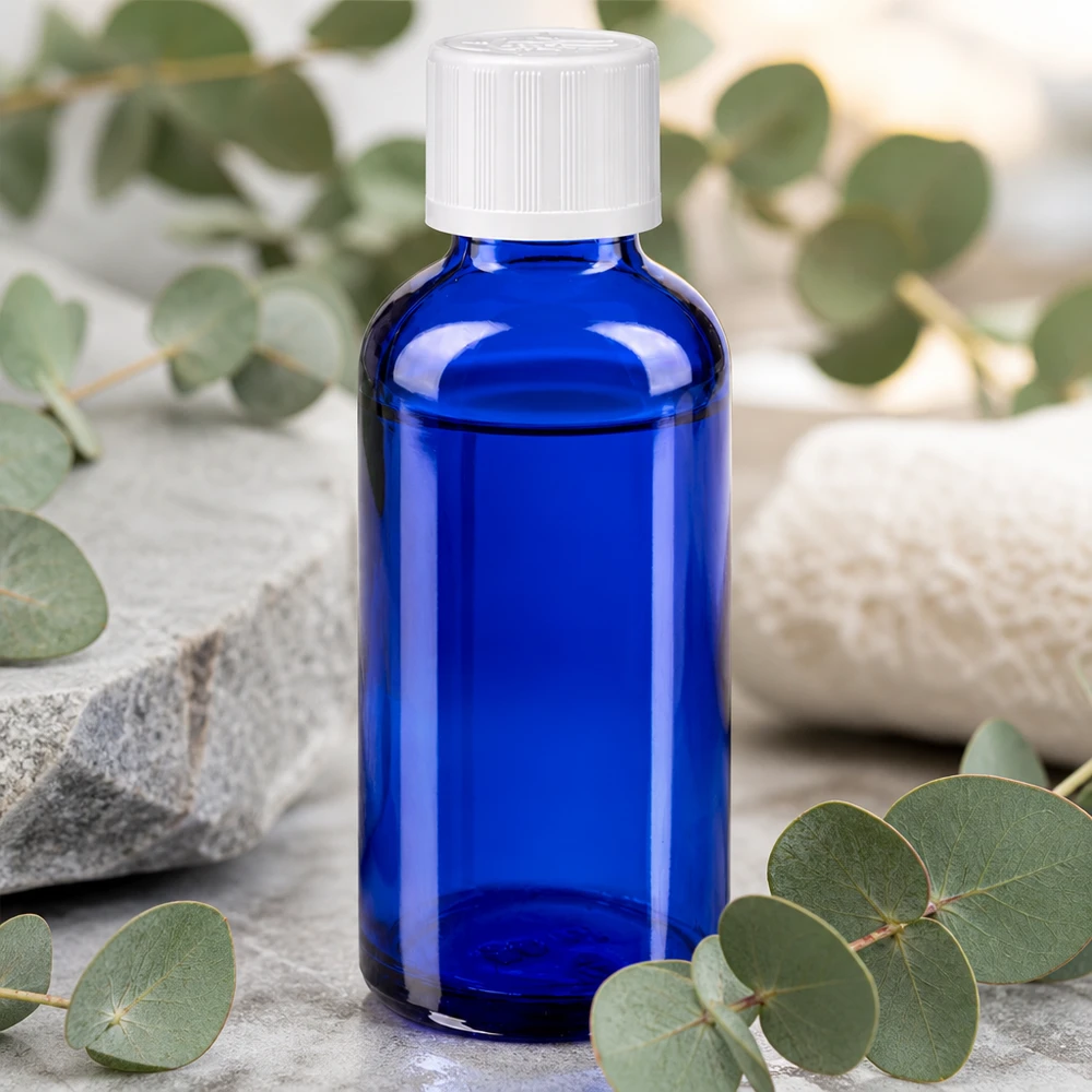 Flacon de pharmacie bleu cobalt de 50 ml muni d'un bouchon KiSi blanc, posé sur une pierre naturelle parmi des branches d'eucalyptus