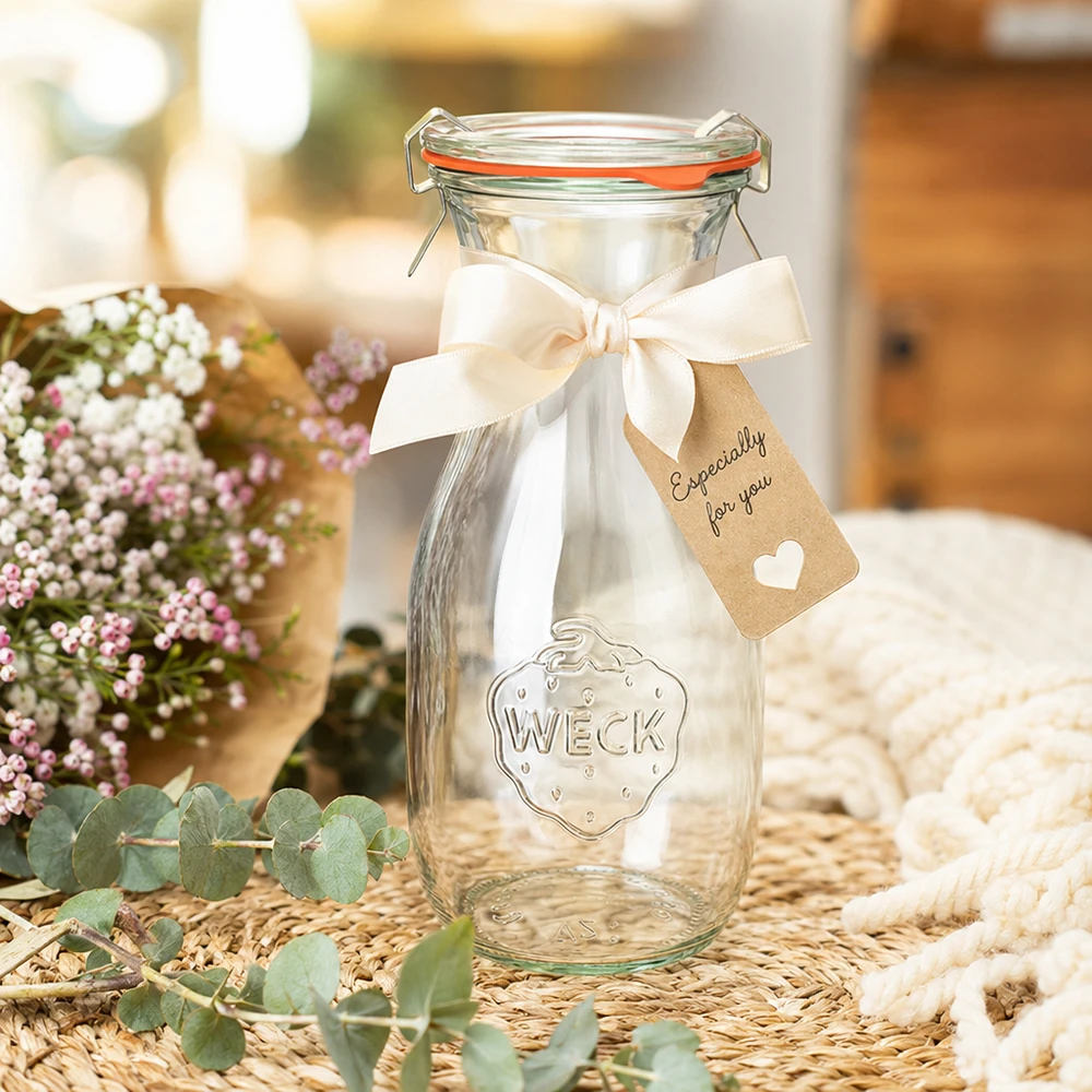 Set completo con bottiglia per succhi da 530 ml WECK RR60 Bottiglia di succo WECK da 530 ml RR60 decorata con un nastro bianco e un'etichetta regalo disposta tra i fiori