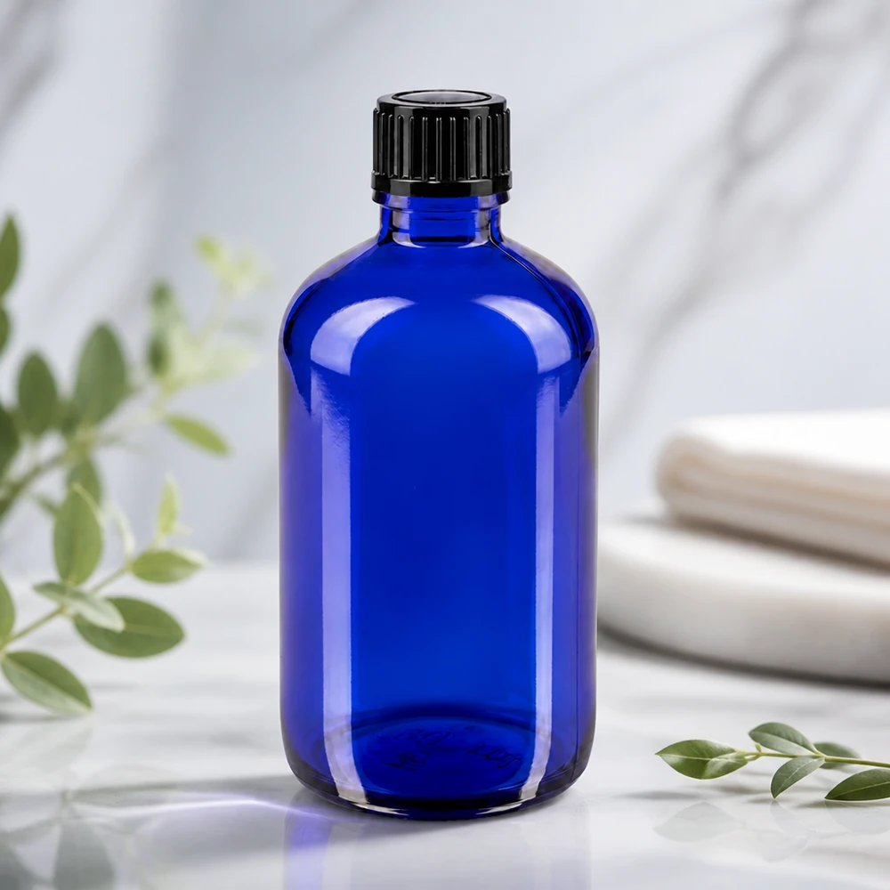 Flacon de pharmacie bleu 100ml bouchon compte-gouttes 1mm noir Std Bouteille en verre bleue de 100 ml avec un bouchon noir strié, posée sur un fond en marbre blanc orné de branches d'eucalyptus