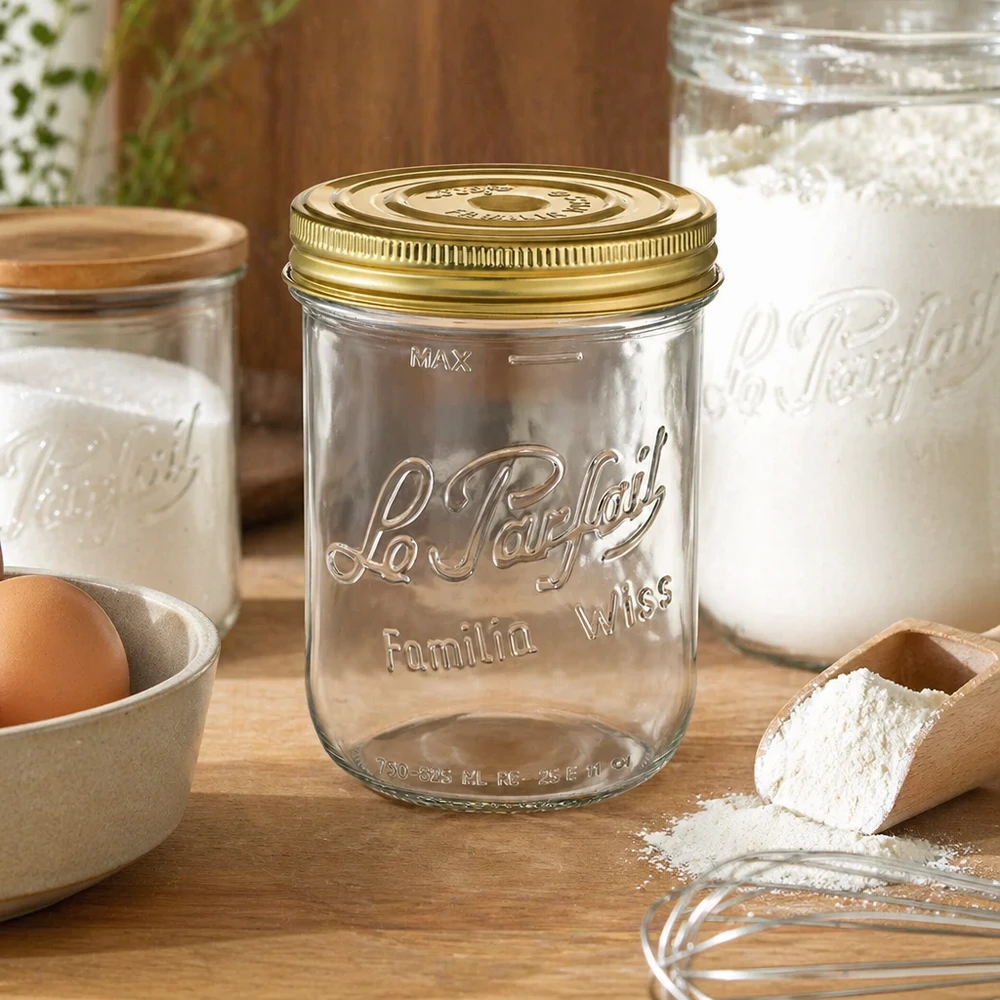 Le Parfait® Familia Wiss 1 kg Ø100 mm vaso Vasetto Familia Wiss da 1020 ml con coperchio dorato sul tavolo di legno accanto a ingredienti da forno e altri vasetti per conserve
