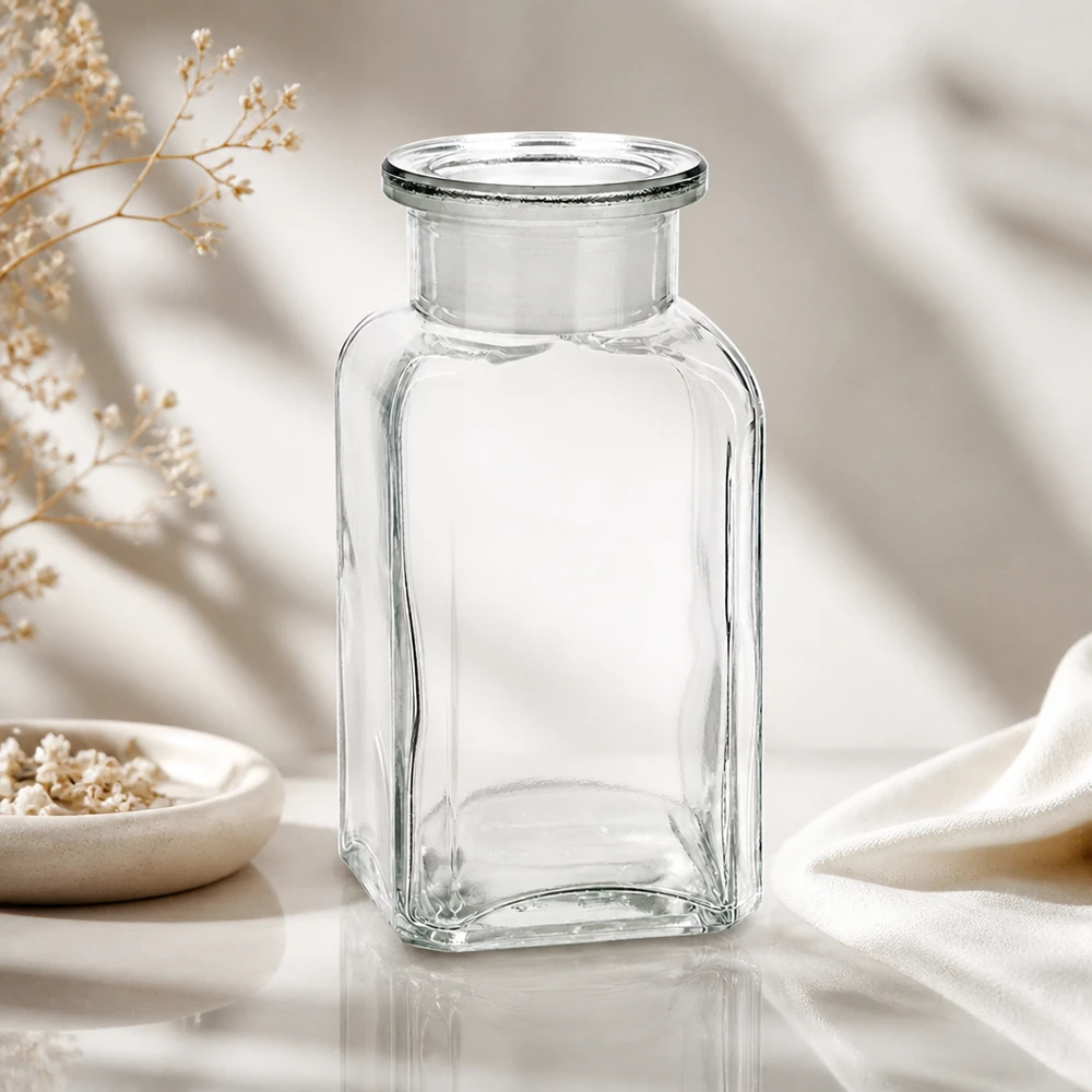 Bottiglia vuota trasparente a forma quadrata da 100 ml con collo largo, su uno sfondo di elegante tessuto bianco e fiori secchi