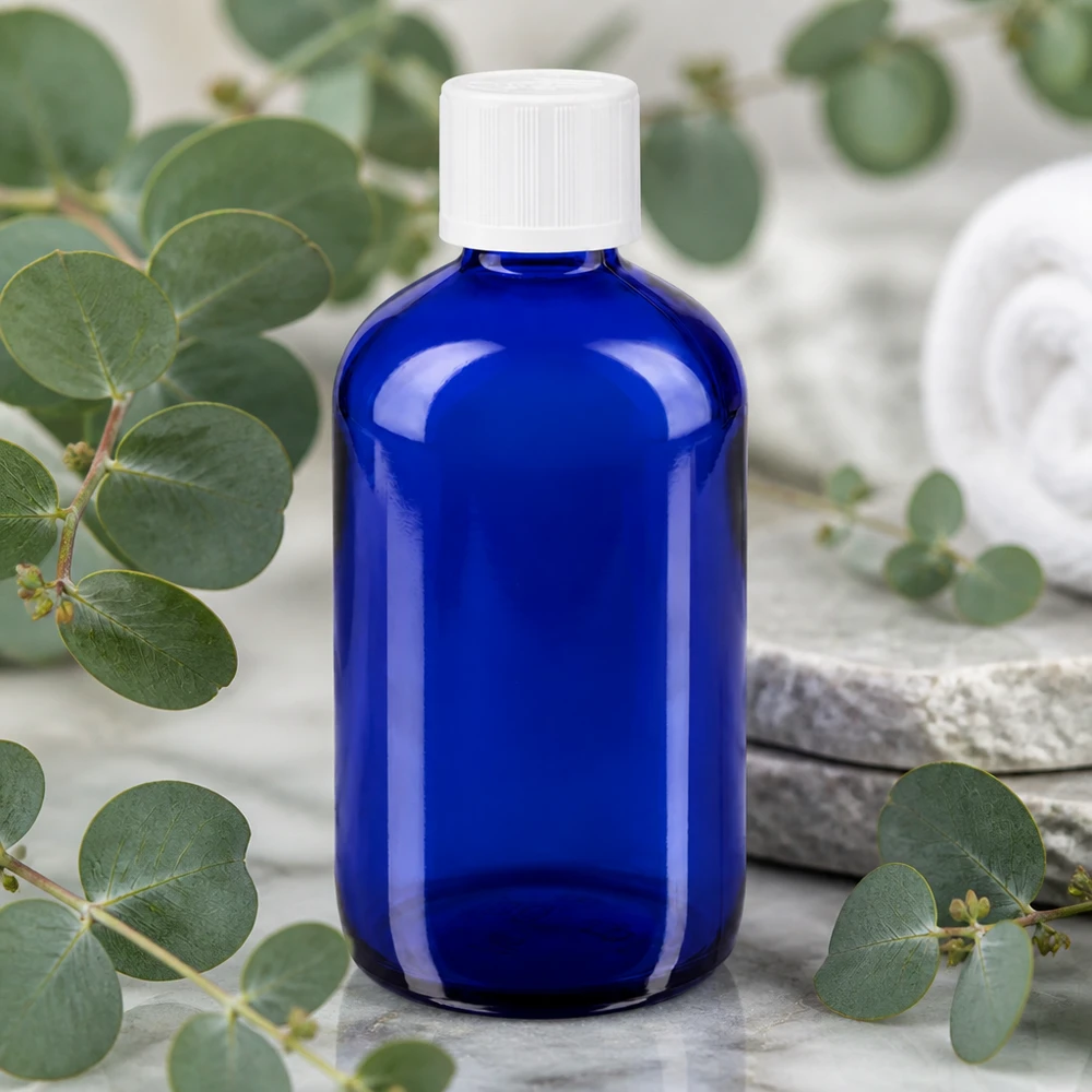 Flacon compte-gouttes bleu de 100 ml en verre d'apothicaire, muni d'un bouchon KiSi blanc, entouré de branches d'eucalyptus