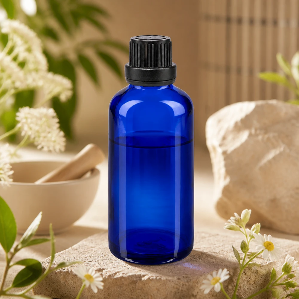 Flacon en verre bleu cobalt de 50 ml avec bouchon à vis noir, posé sur une pierre naturelle ornée de fleurs de camomille et d'un mortier