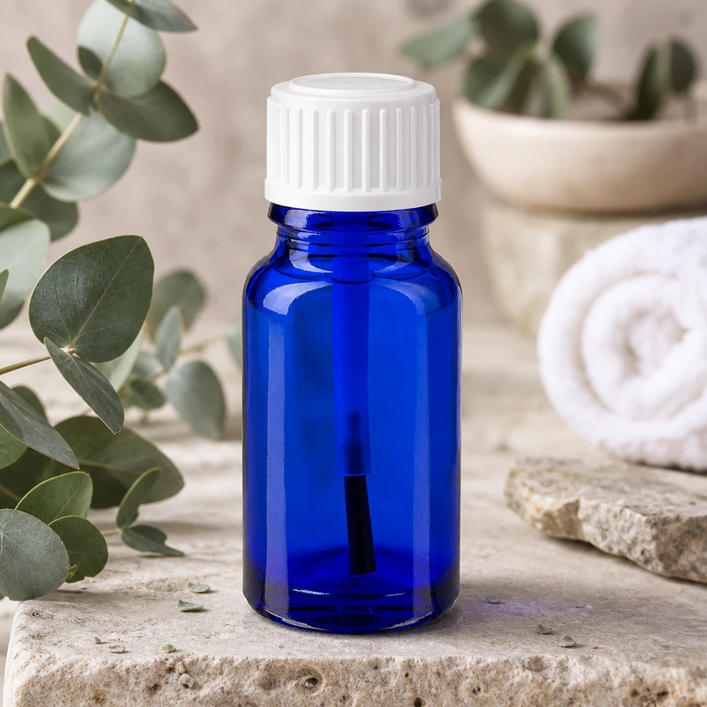 Flacon de pharmacie bleu 10ml bouchon vissable blanc pinceau Flacon en verre bleu de 10 ml muni d'un embout pinceau blanc, posé à côté de branches d'eucalyptus sur une pierre naturelle
